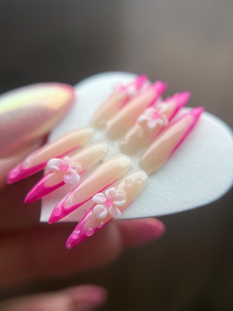 May include: A collection of long, pointed artificial nails. Each nail has a pale pink base, a vibrant pink tip, and a 3D white flower decoration. The nails are displayed on a white heart-shaped surface.