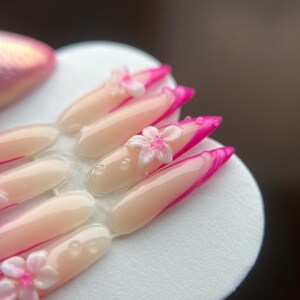May include: A set of artificial nails with a pale pink base and vibrant pink tips. Each nail features a small, white flower with a pink centre. The nails are arranged on a white, circular surface.