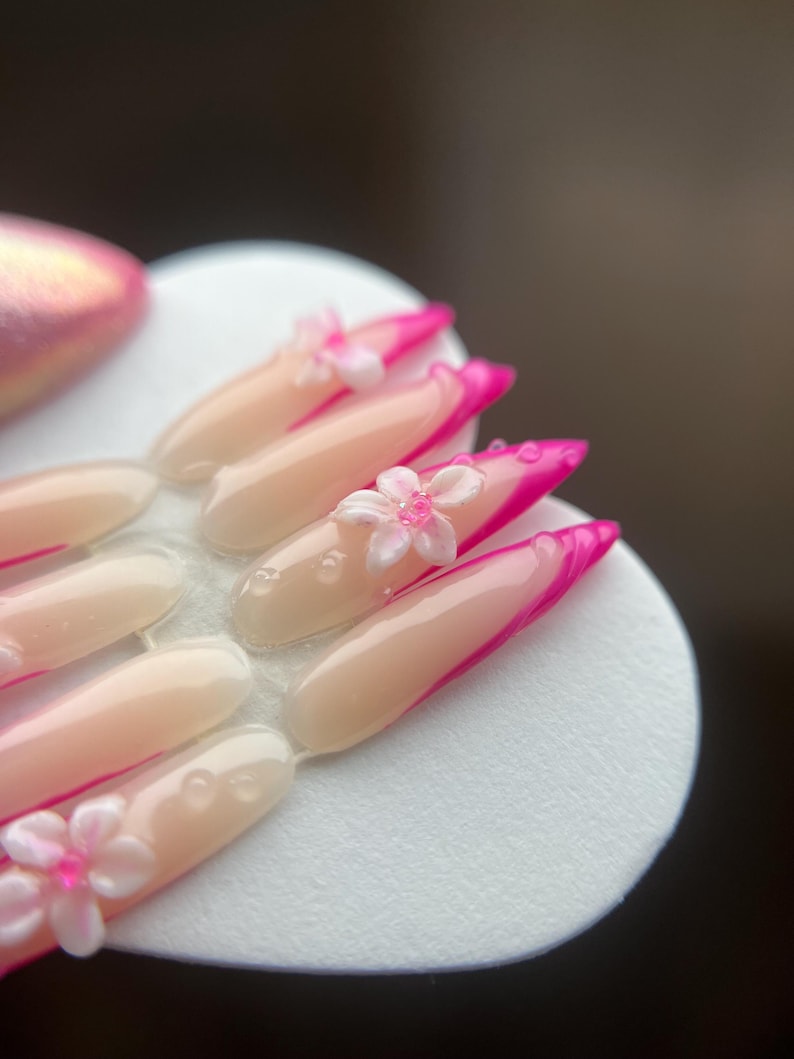 May include: A set of artificial nails with a pale pink base and vibrant pink tips. Each nail features a small, white flower with a pink centre. The nails are arranged on a white, circular surface.