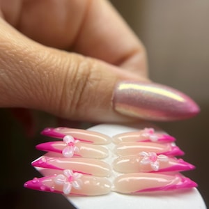 May include: A set of artificial nails with a pink and white design. The nails have a French tip style with a bright pink colour. Each nail is adorned with a small, white flower. The nails are held on a white surface.