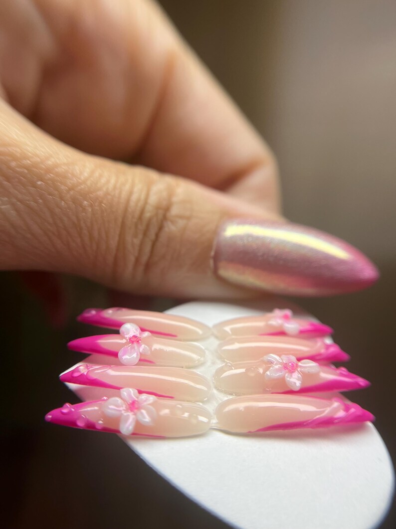 May include: A set of artificial nails with a pink and white design. The nails have a French tip style with a bright pink colour. Each nail is adorned with a small, white flower. The nails are held on a white surface.