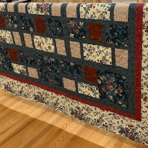 May include: A handmade quilt with a patchwork design. The quilt features a central panel with dark blue and beige floral patterns, accented by red squares and borders. The edges are finished with a floral print on a cream background.
