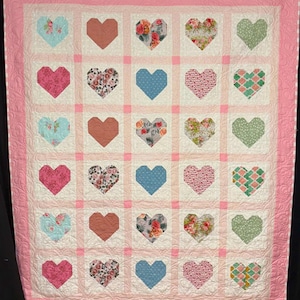 Puede incluir: Una colcha acolchada con un patrón repetido de formas de corazón. Los corazones son de varios colores y estampados, incluyendo estampados florales, rosa liso, azul y verde. La colcha tiene un borde rosa y un fondo blanco.