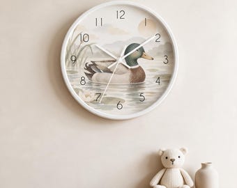 May include: A round wall clock with a white frame and a white face featuring a mallard duck swimming in a pond. The clock has black numbers from 1 to 12, white hour, minute, and second hands, and a background illustration of reeds and a sun.