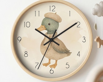 May include: A round wall clock with a light wood frame and a cream-coloured face. The centre features a watercolour illustration of a duck wearing a tan beret and a matching scarf. The clock has black numerals and thin black hour, minute, and second hands.