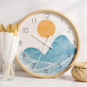 May include: A round wall clock with a light wood frame and a white face. The clock features a graphic of a yellow sun above blue textured mountains and wavy lines. It has black numerals from 1 to 12 and white clock hands indicating the time.