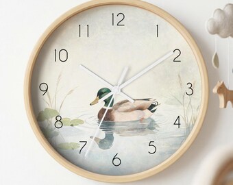 May include: A round wall clock with a light wood frame and a white face. The clock features a watercolour illustration of a mallard duck swimming in water with lily pads and reeds. It has black numerals from 1 to 12 and thin white clock hands.