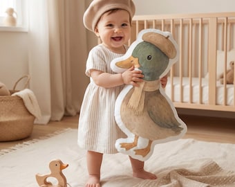 May include: A plush cushion shaped like a duck wearing a tan beret and a matching scarf. The duck has a green head, brown body, and orange feet. The cushion features a soft, white border and a printed illustration of the duck in a neutral colour palette.