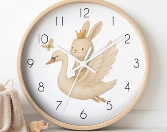 May include: A round wall clock with a light wood frame and a white face featuring black numbers from 1 to 12. The central illustration depicts a beige rabbit wearing a small gold crown, riding on the back of a white swan, with a small butterfly nearby.