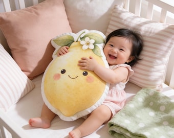 May include: A plush lemon-shaped cushion featuring a printed smiling face with rosy cheeks, green leaves, and a small white flower at the top. The cushion has a white border and a soft, textured surface, designed in a bright yellow colour palette.