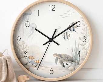 May include: A round wall clock with a light wood frame and a white face featuring a watercolour illustration of an underwater scene. The design includes a sea turtle, jellyfish, coral, and small fish with black numerals and black clock hands.