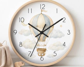 May include: A round wall clock with a light wood frame and a white face featuring a watercolour illustration of a hot air balloon, clouds, a crescent moon, and a small star. The clock has black hour, minute, and second hands and black numerals.