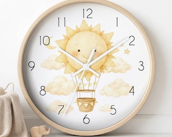 May include: A round wall clock with a light wood frame and a white face. The centre features a watercolour illustration of a smiling sun in a hot air balloon floating among clouds, stars, and a crescent moon. It has black numbers and white hands.