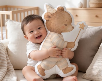 May include: A white shaped throw pillow featuring a watercolour illustration of a brown bear wearing a party hat and striped pyjamas, holding a star-tipped wand. The bear has closed eyes and is depicted in a standing position on the pillow.