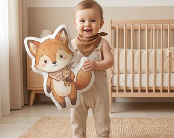 May include: A plush throw cushion shaped like a cartoon fox with orange and white fur, wearing a small brown scarf and holding a tiny teddy bear. The fox has large eyes, a dark nose, and a bushy tail, rendered in a soft, illustrated style.