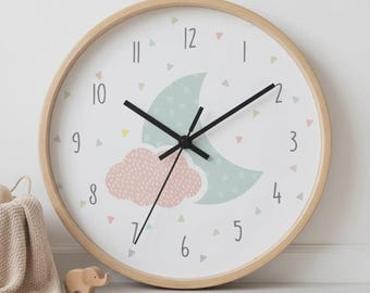 May include: A round wall clock with a light wood frame and a white face. The clock features black hands, dark grey numbers, and a central graphic of a light green crescent moon with a small pink cloud, surrounded by tiny colourful triangles.