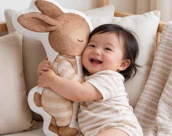 May include: A plush cushion shaped like a standing rabbit with brown fur and a beige striped outfit. The rabbit has closed eyes and a simple facial expression. The cushion is white-edged and features a soft, watercolour-style illustration design.
