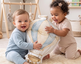 Op de afbeelding: Een pluche kussen in de vorm van een heteluchtballon met een aquarelontwerp in blauw- en beigetinten, voorzien van een gedetailleerde illustratie van de mand en ballon, vastgehouden door twee baby's op een zacht, crèmekleurig tapijt.