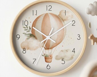 May include: A round wall clock with a light wood frame and a cream-coloured face. The clock features a central illustration of a beige and white striped hot air balloon with a basket, surrounded by watercolour clouds, green leaves, and stars.