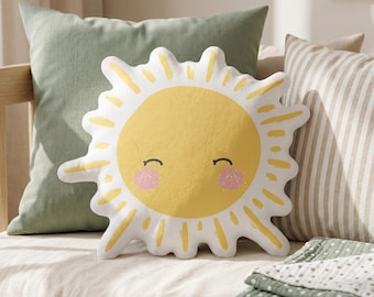 May include: A sun-shaped throw cushion with a white background and a yellow centre. The sun features a smiling face with closed eyes and pink cheeks with white speckles. The rays are yellow and white, extending outwards in a jagged, irregular pattern.