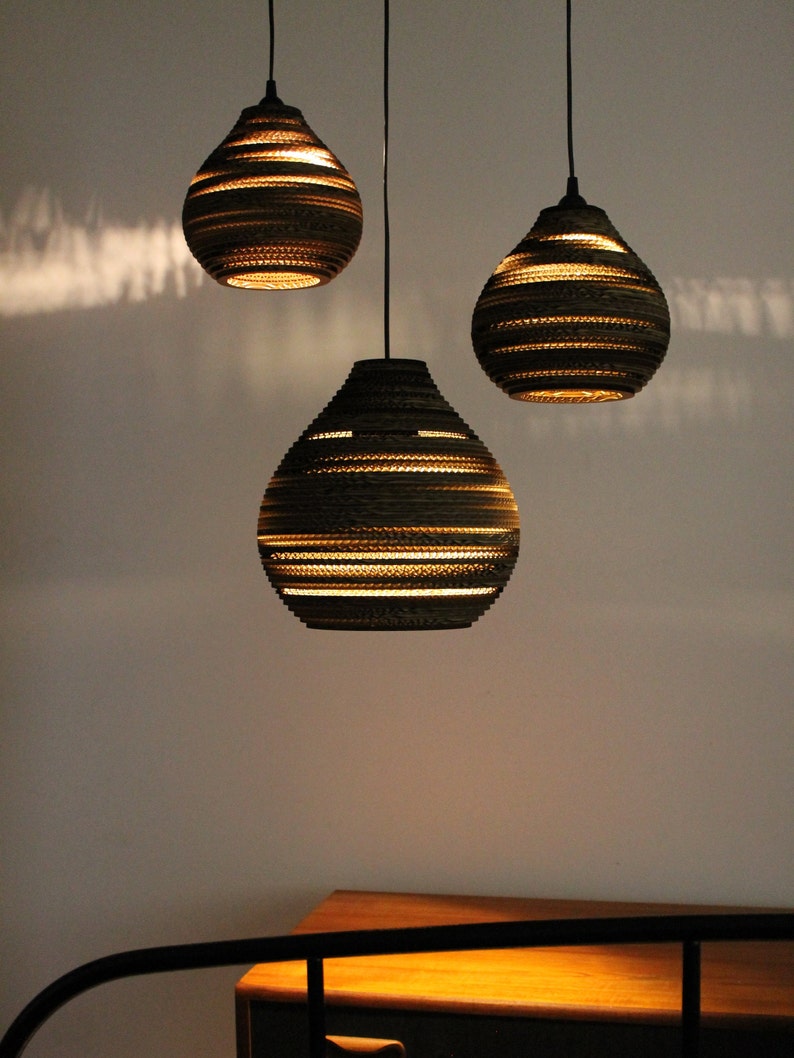 May include: Three hanging pendant lights made of cardboard with a layered design. The lights are turned on and emit a warm glow.