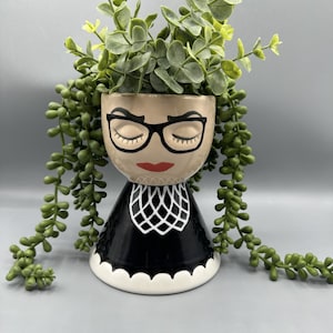 May include: A ceramic planter shaped like a woman's face with closed eyes, black glasses, and red lips. The planter is black with a white collar and a white scalloped edge. The planter is filled with green plants.