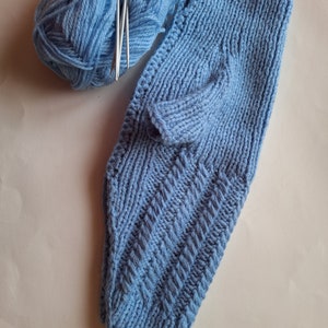 May include: A blue knitted baby garment with a button on the shoulder. The garment is lying on a white surface next to a ball of blue yarn and knitting needles.