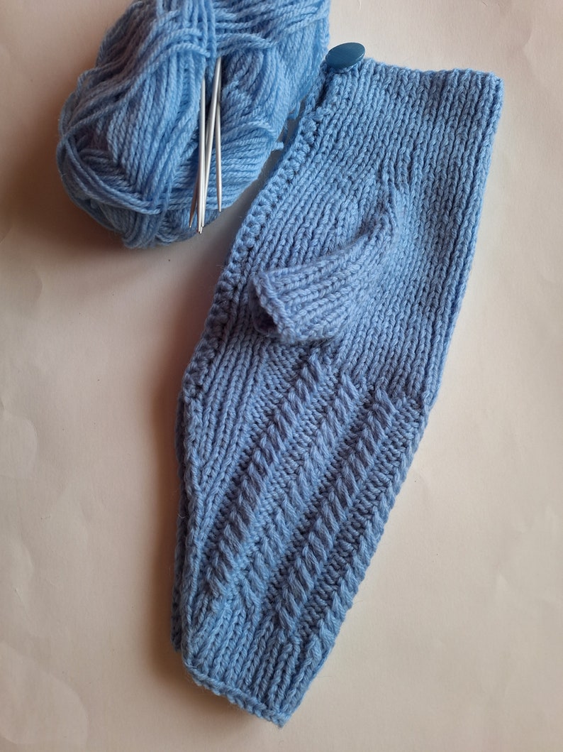 May include: A blue knitted baby garment with a button on the shoulder. The garment is lying on a white surface next to a ball of blue yarn and knitting needles.