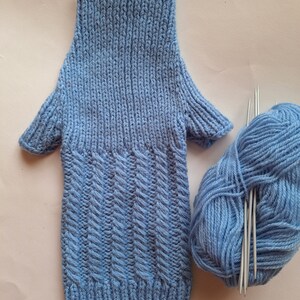 May include: A light blue knitted fingerless glove with a textured pattern. The glove is lying on a white surface next to a ball of light blue yarn and knitting needles.