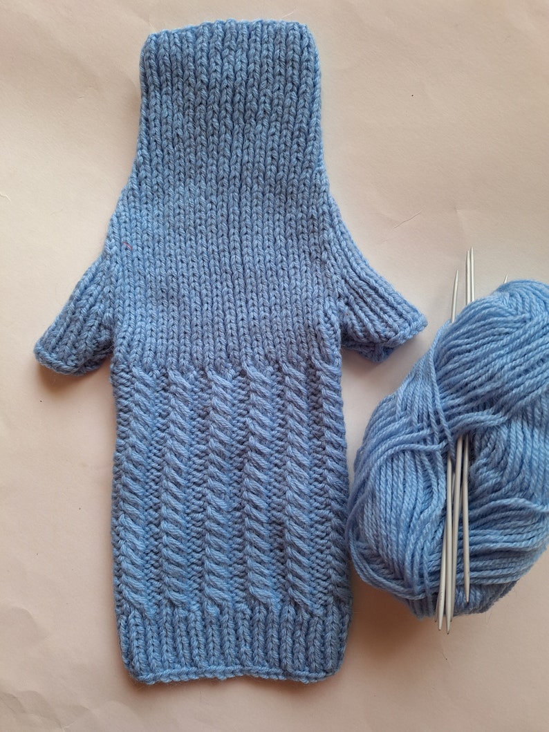 May include: A light blue knitted fingerless glove with a textured pattern. The glove is lying on a white surface next to a ball of light blue yarn and knitting needles.