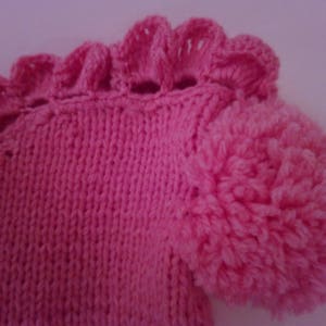 May include: A pink knitted baby hat with a fluffy pom-pom on top. The hat has a ruffled edge.