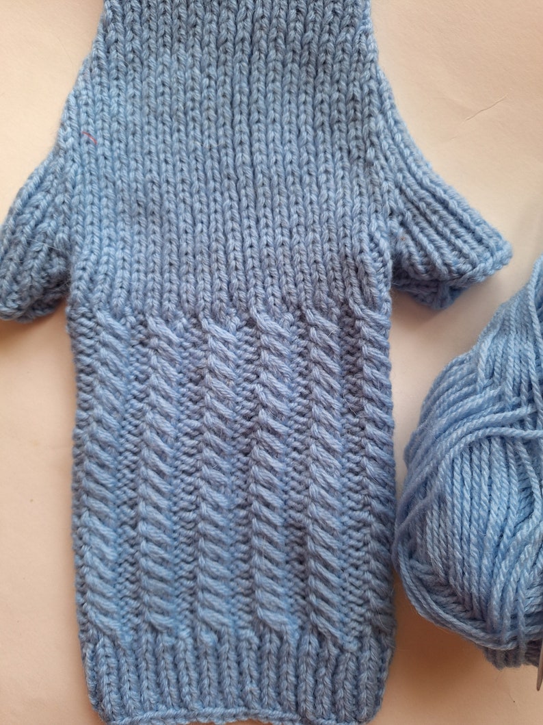 May include: A light blue knitted garment with a textured pattern. The garment has long sleeves and a ribbed hem. A ball of light blue yarn is also pictured.
