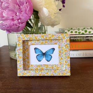 May include: A small picture frame with a yellow and white floral pattern, displaying a blue butterfly image. The frame rests on a wooden surface. A vase of pink and white flowers is visible in the background.