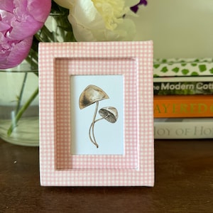 May include: A small, pink gingham-patterned picture frame with a mushroom illustration. The frame holds a print of two brown and tan mushrooms. The frame is sitting on a dark brown surface, with flowers in the background.