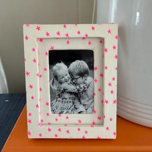 May include: A rectangular picture frame with a matte off-white finish decorated with small, scattered neon pink stars. The frame holds a black and white photograph of two children embracing, with the child on the left wearing a patterned dress.
