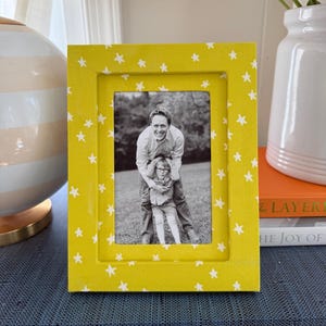 May include: A rectangular picture frame with a bright yellow finish and a repeating pattern of small white stars. The frame holds a black and white photograph of an adult standing behind a child, both smiling, set against an outdoor background.