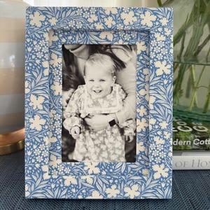 Op de afbeelding: Een rechthoekige fotolijst met een blauw-wit bloemenpatroon. De lijst bevat een zwart-witfoto van een lachend kind dat door een volwassene wordt vastgehouden. De lijst heeft een platte, brede rand met een herhalend botanisch ontwerp.