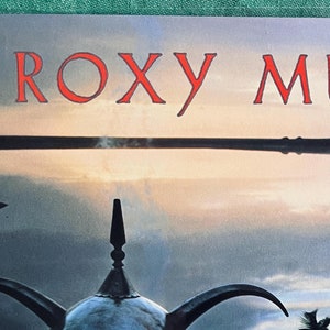 May include: A vintage album cover featuring a still life image of a silver teapot with a long spout and a handle. The background is a hazy, blue sky with a reflection of the teapot in a body of water. The album title "ROXY MUSIC" is printed in red at the top of the cover.
