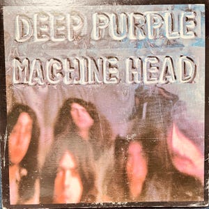 May include: Album cover for Deep Purple's Machine Head. The album title is embossed in a silver, textured font at the top. The band members are visible in the lower portion of the cover, with a blurred, purple and pink background.