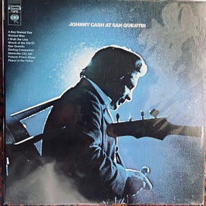 May include: A vintage vinyl record cover featuring a black and white photo of Johnny Cash playing a guitar. The album title "JOHNNY CASH AT SAN QUENTIN" is printed in white letters on a blue background. The tracklist includes "A Boy Named Sue", "Wanted Man", "I Walk the Line", "Wreck of the Old 97", "San Quentin", "Darling Companion", "Starkville City Jail", "Folsom Prison Blues", and "Peace in the Valley".