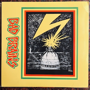 May include: A yellow, green and red framed image of a black and white illustration of a lightning bolt striking a building with a dome. The text "BAD BRAINS" is written vertically in red and green on the left side of the image.