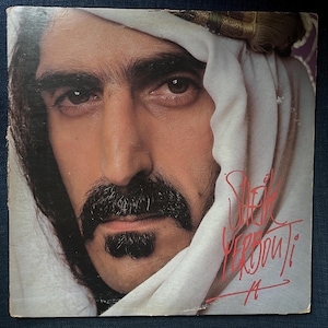 Peut inclure: Pochette d'album avec un gros plan d'un homme avec une moustache et une barbe sombres, portant un headdress blanc. Le titre de l'album "Sheik Yerbouti" est écrit en script rouge.