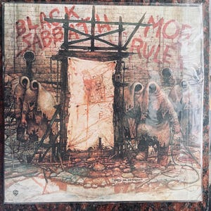 May include: Black Sabbath album cover art featuring a brick wall with the words "Black Sabbath" and "Mob Rules" painted on it.  Three hooded figures stand in front of a white sheet with red splatter paint. The text "Greg Hildebrandt" is visible at the bottom of the image.
