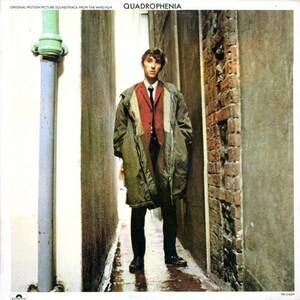May include: A man in a green coat, red vest, and black pants stands in a narrow alleyway. The alleyway is lined with brick walls and a white wall with a pipe running down it. The man is looking at the camera. The image is from the album cover for the film Quadrophenia.