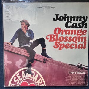 May include: A vinyl record album cover for Johnny Cash's "Orange Blossom Special." The cover features a photograph of Johnny Cash sitting on a train car. The album title is in large, red, stylized lettering. The album's tracklist is listed on the bottom right.