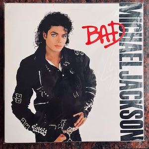 May include: A vinyl record album cover featuring Michael Jackson in a black leather jacket with silver studs. The album title "BAD" is written in red letters. The background is white.