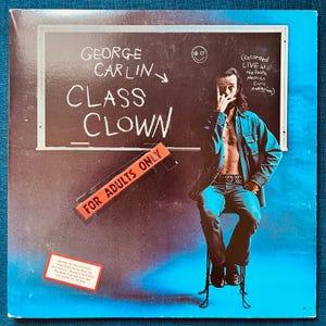 May include: Album cover for George Carlin's "Class Clown." The cover features a chalkboard with the title and the words "For Adults Only." A man is seated in front of the chalkboard. The album cover is on a blue background.