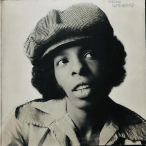 May include: Black and white portrait of a person wearing a striped newsboy cap and a collared shirt. The name "KEVIN HUTCHINGS" is written in blue ink at the top right corner of the image.