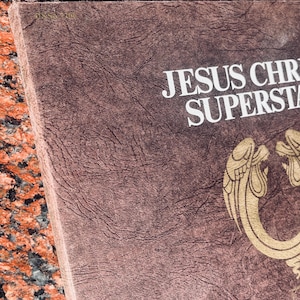 May include: A brown vinyl record album with the title "JESUS CHRIST SUPERSTAR" in white text. The album cover features a gold design of two angels with wings.