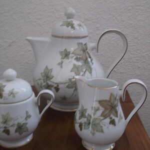 Puede incluir: Un juego de té de porcelana blanca con tetera, azucarero y jarrita para la leche. El juego está decorado con un diseño de hojas verdes y beige y ribetes dorados. La tetera tiene un asa curvada y una tapa con un pequeño pomo.